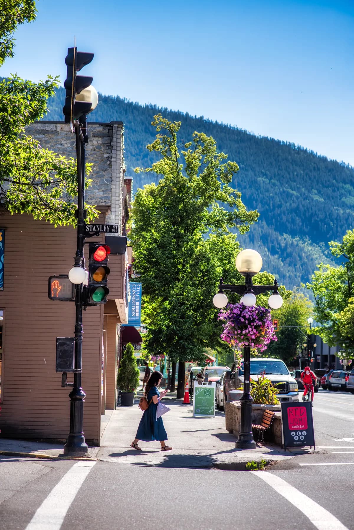 A person crossing a sunny Nelson street with mountain views, trees and hanging flowers